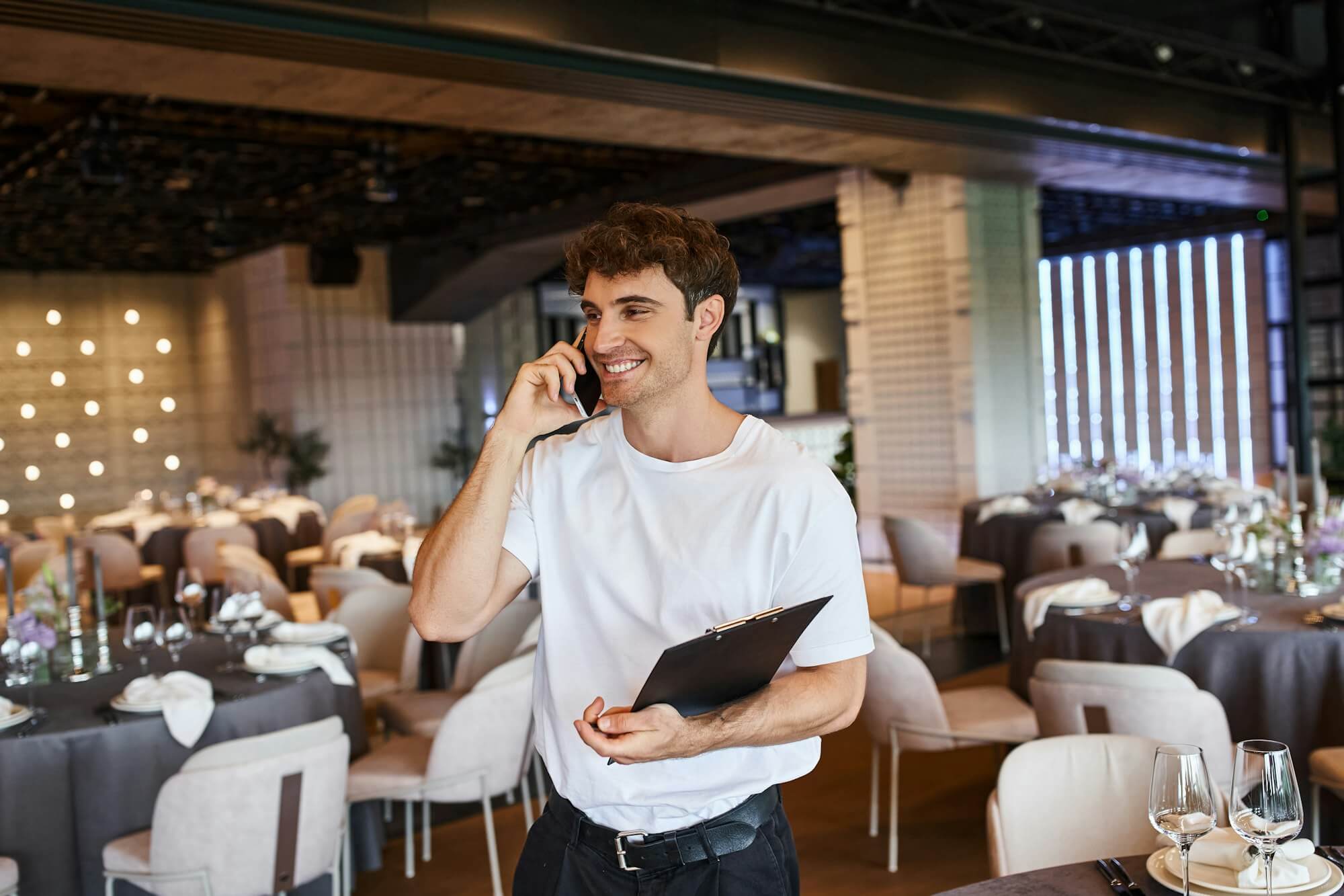 smiling-event-organizer-with-clipboard-talking-on-smartphone-near-festive-tables-in-banquet-hall.jpg